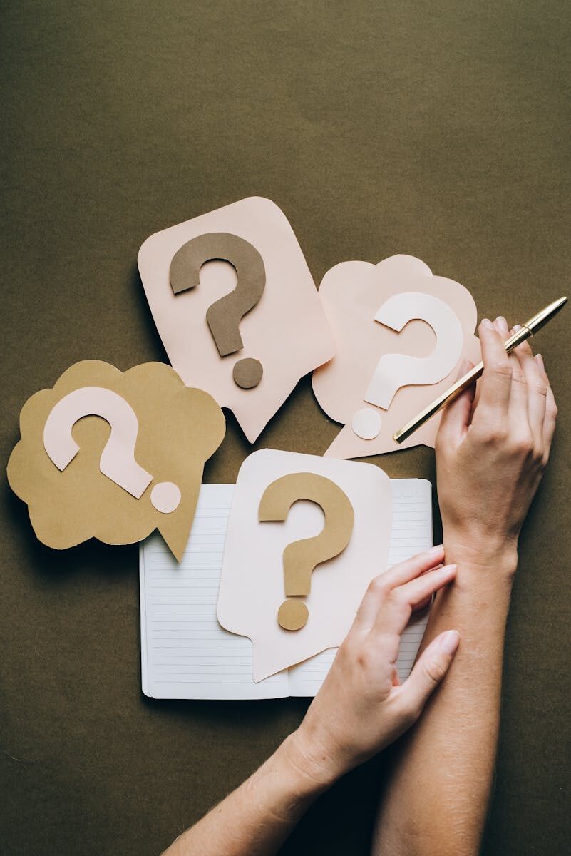 Photo by Leeloo The First Hands holding pen next to paper question marks on notebook, symbolizing inquiry and creativity.